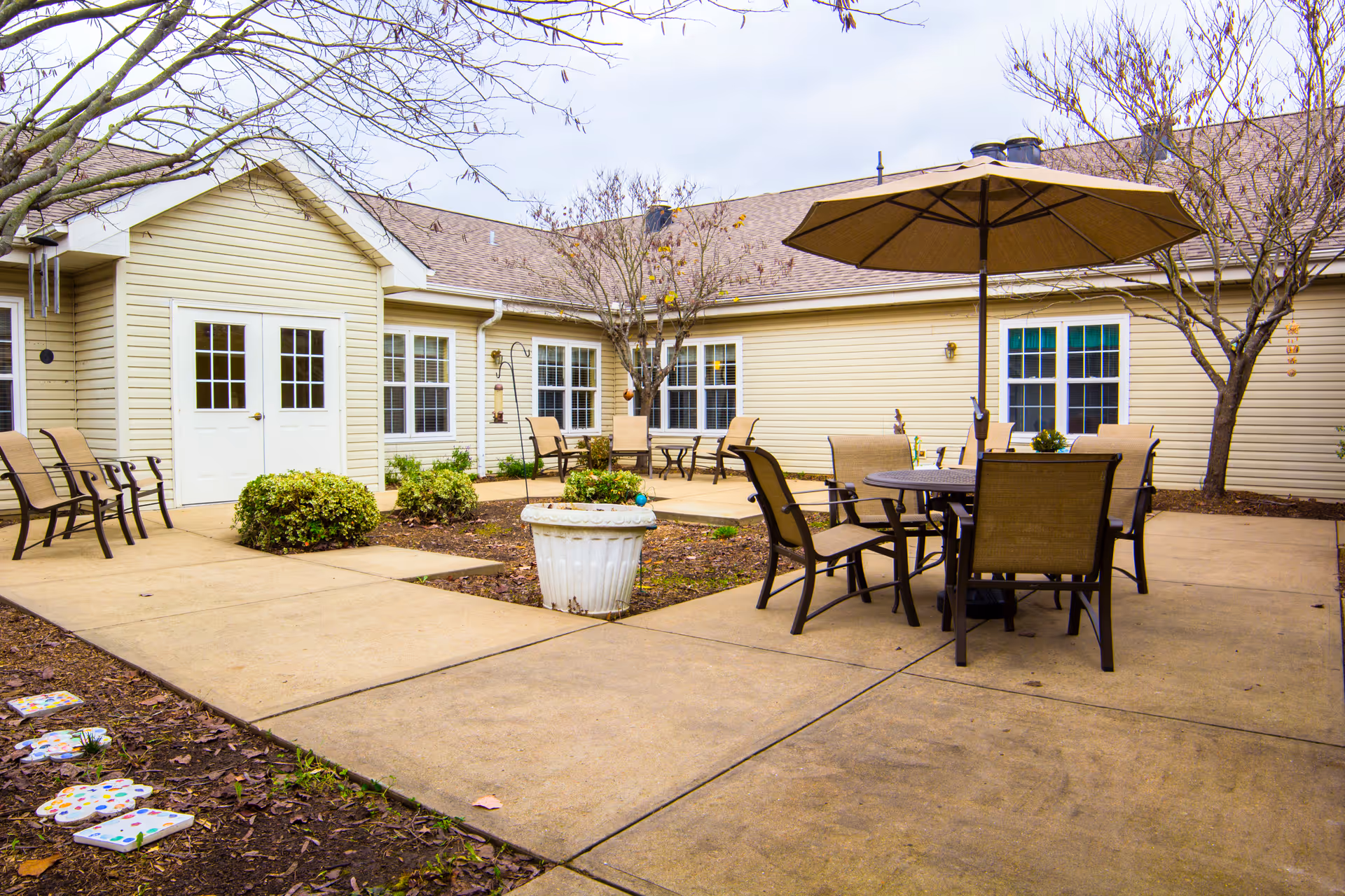 Outdoor courtyard area of a senior living facility with beige siding buildings surrounding a paved patio. The patio has a round table with an umbrella and several chairs, additional benches along the walls, some small trees without leaves, and a large white planter in the center.