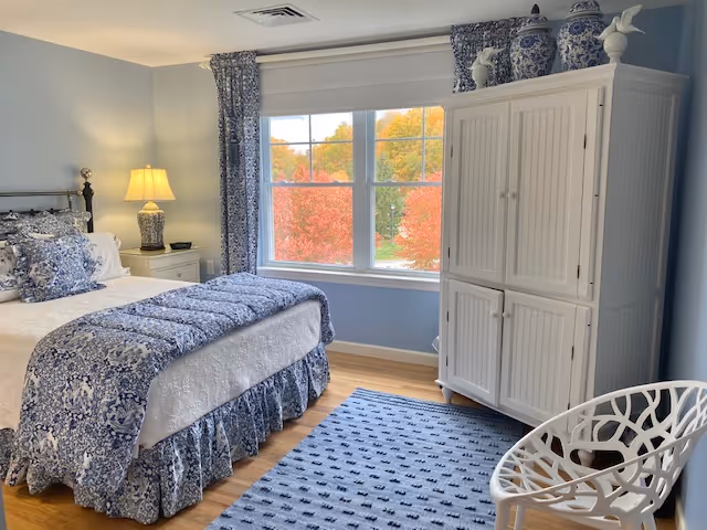 Bright bedroom with blue-and-white patterned bedding, a white armoire, a lamp on a nightstand, and a window showing colorful autumn trees.