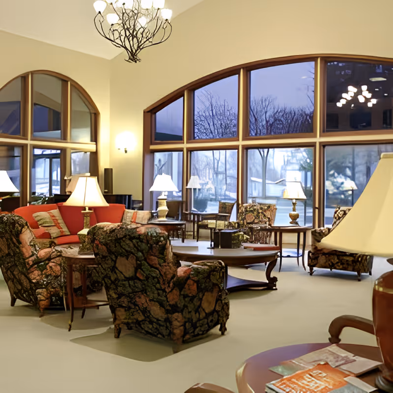 A spacious living room with large arched windows showing a view of trees outside. The room features patterned armchairs, a red sofa, several table lamps, and wooden coffee tables arranged in a cozy seating area. A chandelier hangs from the ceiling, and the room is warmly lit.