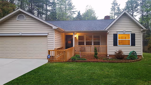 Front exterior view of a single-story house with beige siding, a two-car garage, a small porch with wooden railing, a lit porch light, and a well-maintained lawn and garden area.
