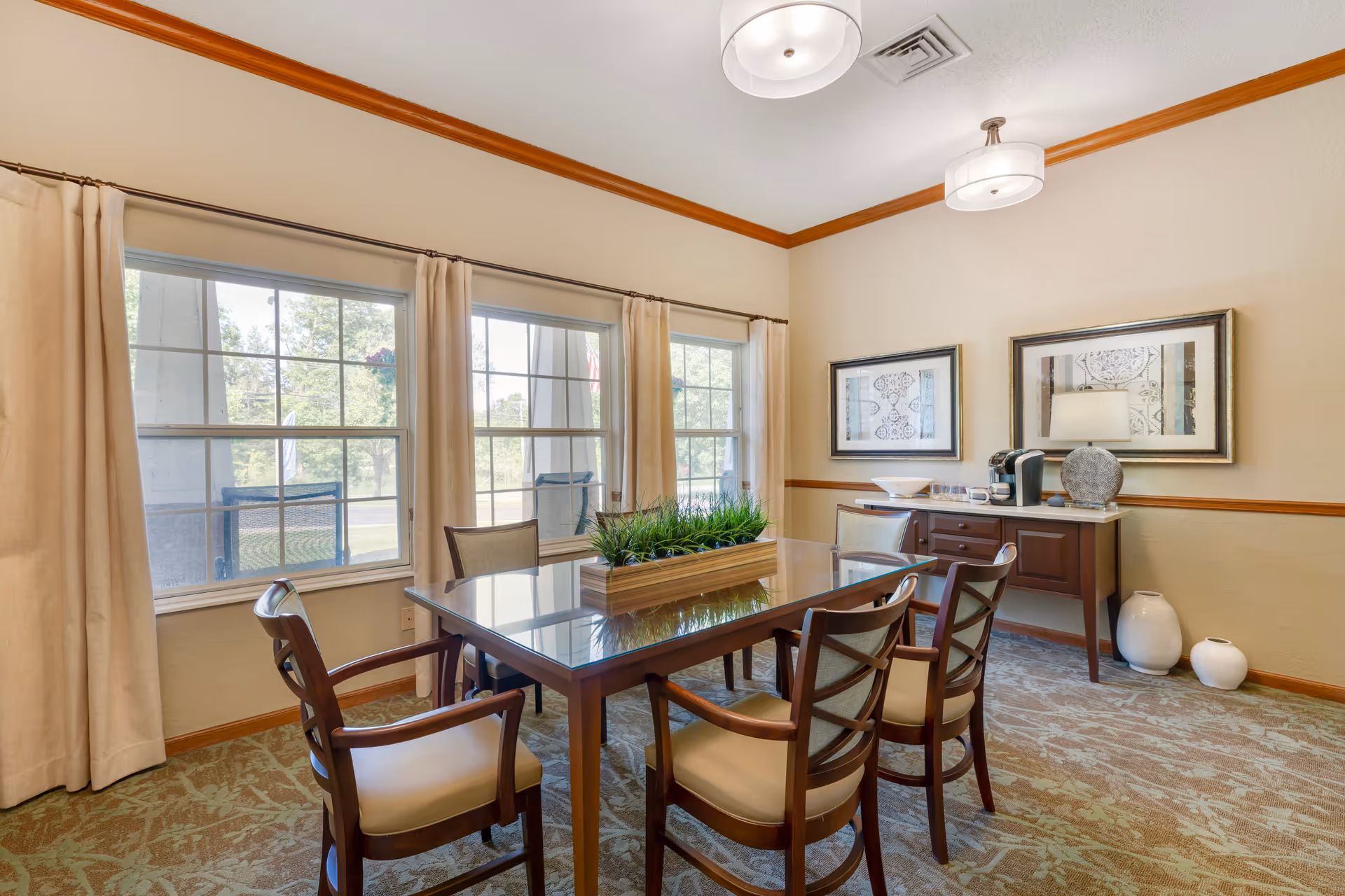 A bright dining room with a glass-top table surrounded by six wooden chairs with cushioned seats. The room has three large windows with beige curtains, a sideboard with a coffee maker and decorative items, two framed artworks on the wall, and two ceiling lights. The floor is carpeted with a patterned design.