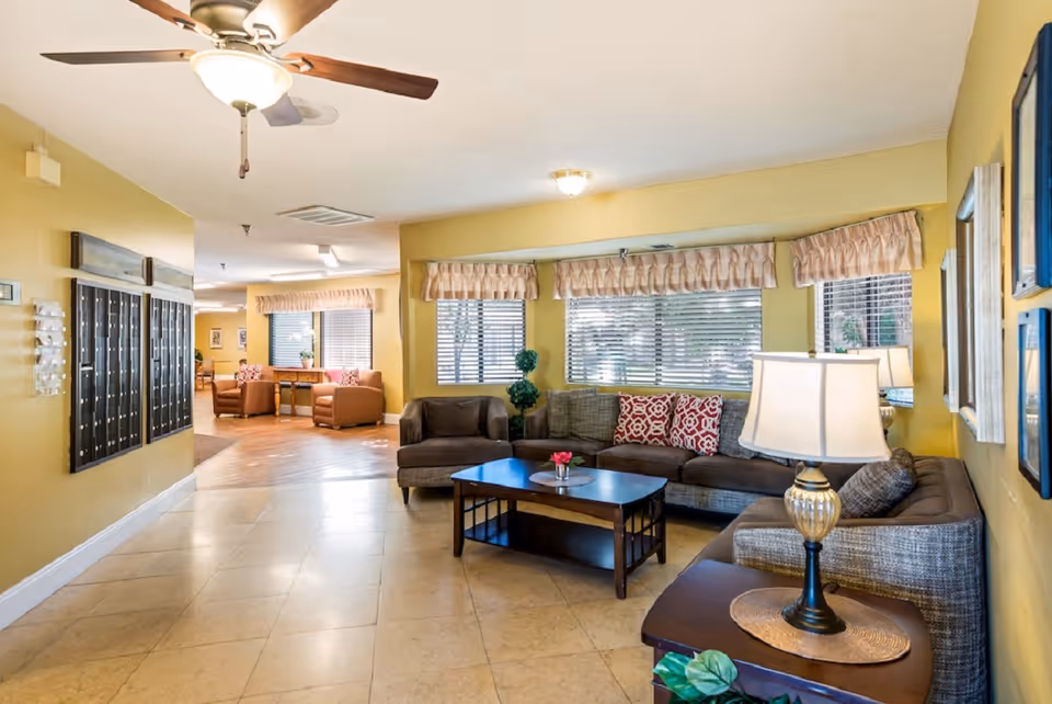 A cozy living room area in a senior living facility with a sectional sofa adorned with patterned cushions, a wooden coffee table with a small flower arrangement, and a side table with a decorative lamp. The room has yellow walls, large windows with blinds and valances, and a ceiling fan with a light fixture. In the background, there is a hallway with additional seating and mailboxes mounted on the wall.