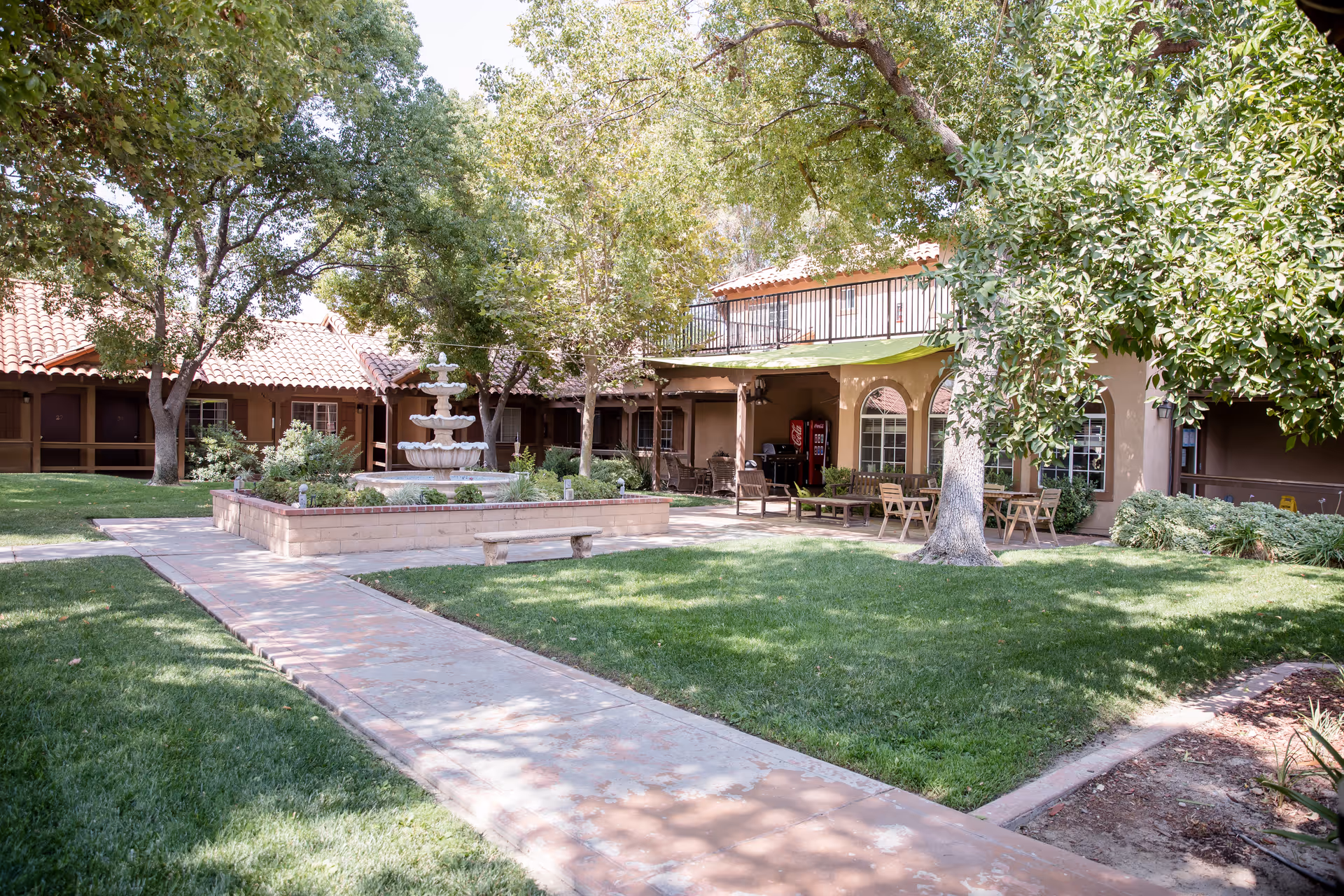 Outdoor courtyard area of a senior living facility with a central multi-tiered fountain surrounded by a raised brick planter. There are trees providing shade, green grass, paved walkways, benches, and outdoor seating with tables and chairs under a covered patio. The building has a tiled roof and arched windows.
