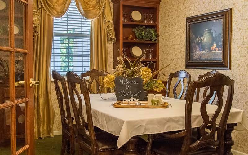 A formal dining room with a table, wooden chairs, floral centerpiece and a small chalkboard that reads 'Welcome to Bickford'.
