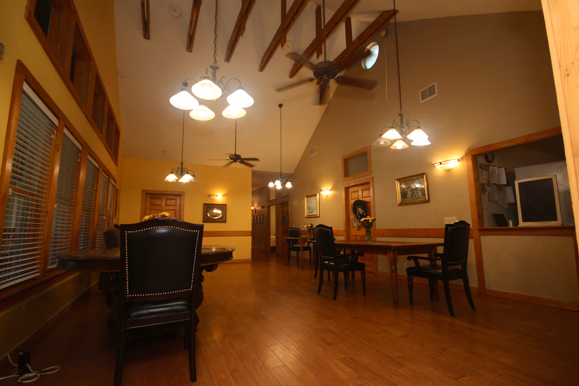 Interior view of a dining area with wooden floors, multiple tables and chairs, ceiling fans, hanging light fixtures, and large windows with blinds on the left side. The walls are decorated with framed pictures and there is a service window on the right.