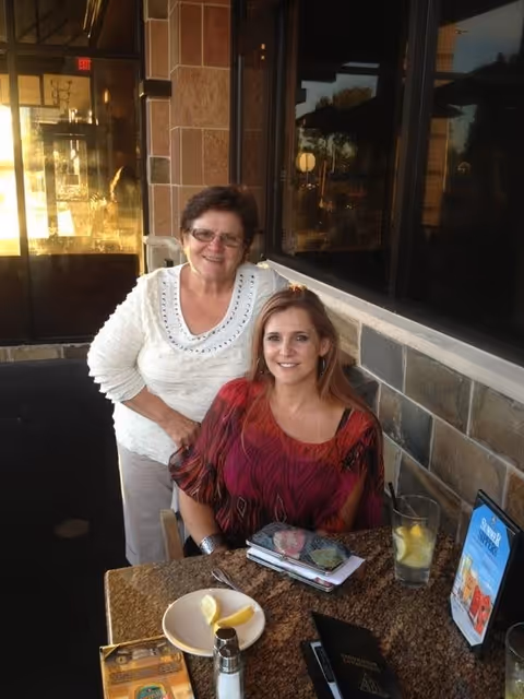 Two women smiling at the camera, one standing and one sitting at a table outside a building with stone walls and large windows. The table has a plate with lemon wedges, a salt or pepper shaker, a glass of water with lemon, and a menu or promotional card.