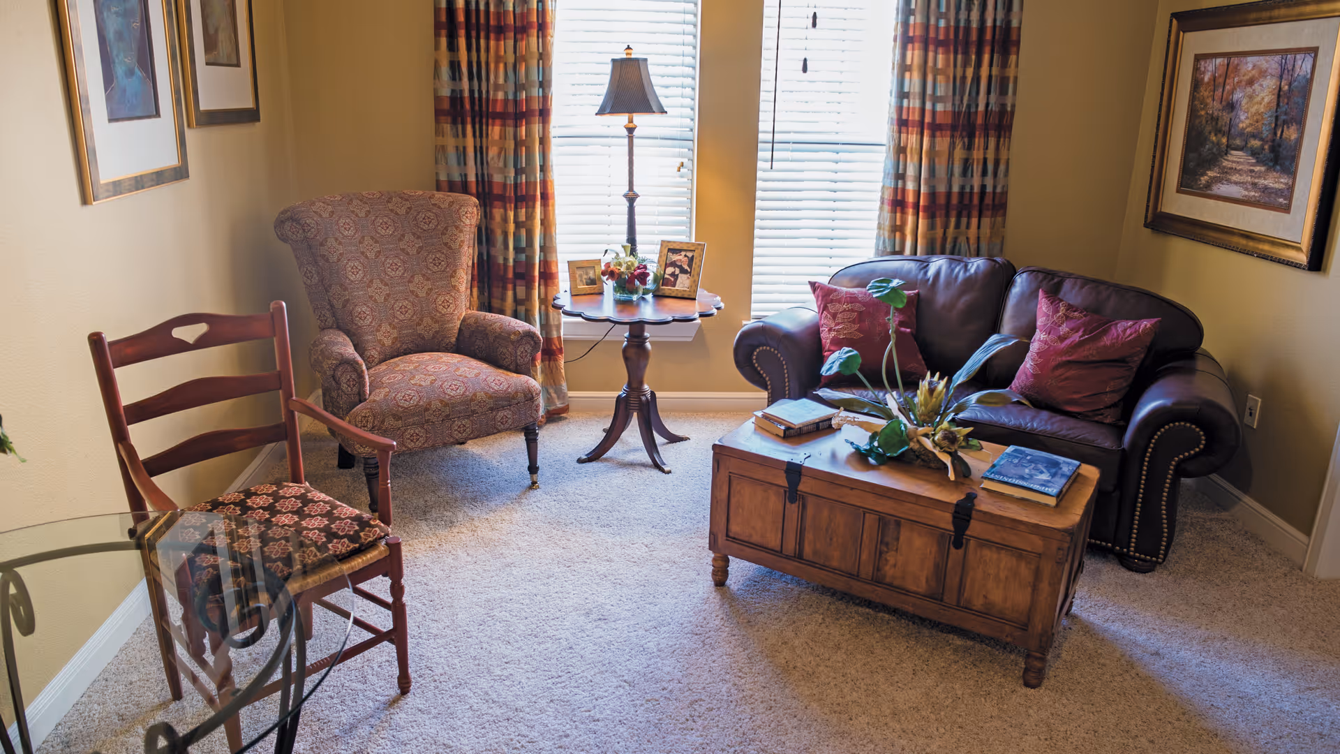 Cozy living room with a leather sofa, patterned armchair, wooden coffee table, side table with a lamp, and a chair by the window.