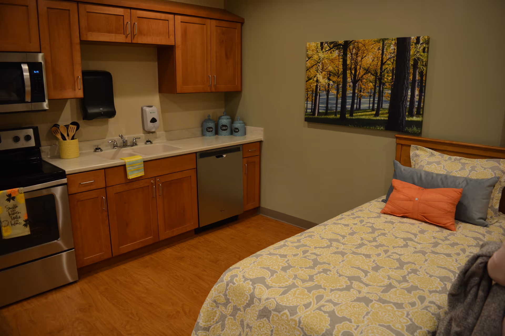 Studio-style room with a bed in the foreground and a kitchenette with wooden cabinets and stainless appliances along the wall.