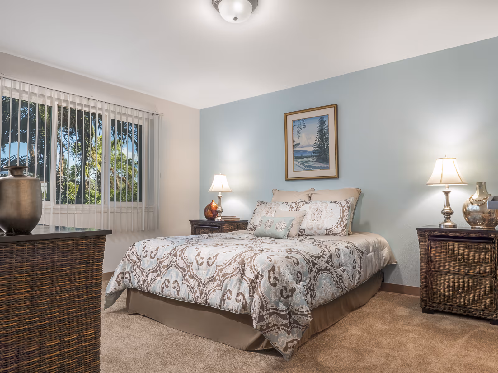 Bedroom with a patterned bed, two nightstands with lamps, a wicker dresser, and a window with vertical blinds.