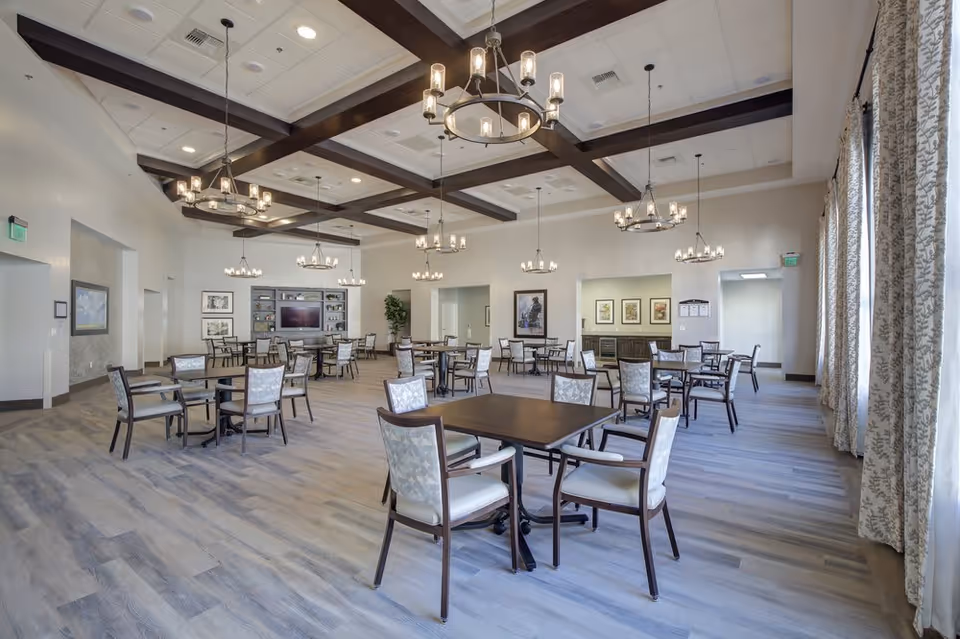 A spacious dining room with multiple square tables and cushioned chairs arranged neatly. The room features a high ceiling with exposed wooden beams and several modern chandeliers hanging down. Large windows with patterned curtains allow natural light to fill the space. The walls are decorated with framed artwork, and there is a cabinet with a television and decorative items at the far end of the room.