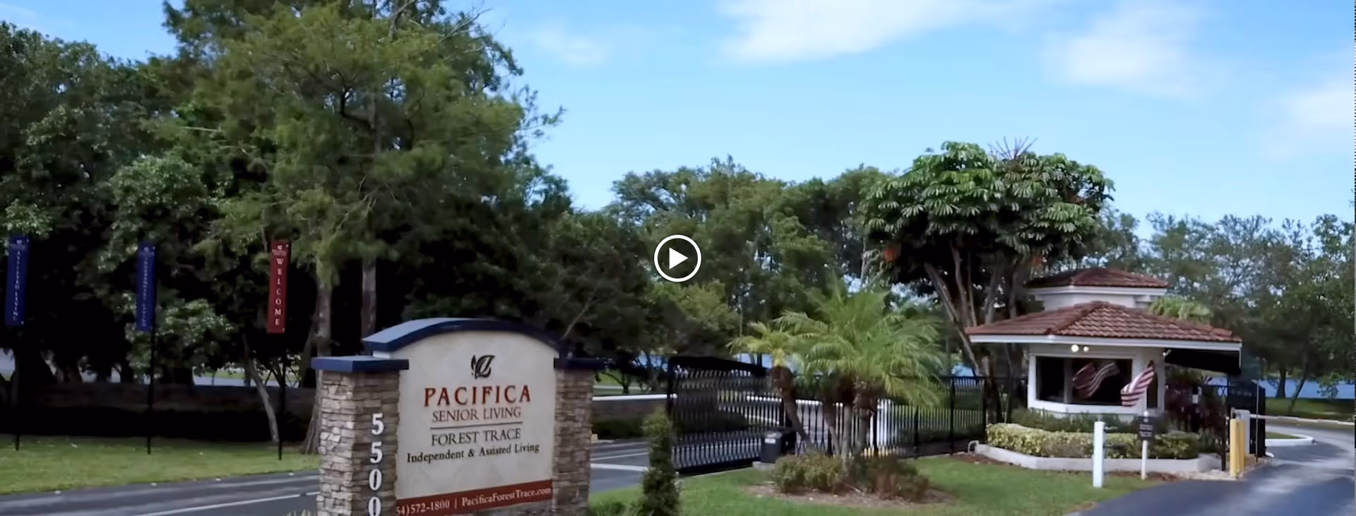 Entrance to Pacifica Senior Living Forest Trace facility with a stone sign displaying the name and contact information, a gated driveway, and a guardhouse surrounded by trees and landscaping under a blue sky.