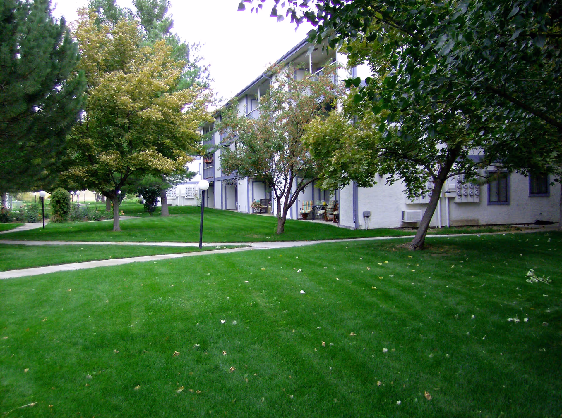 Outdoor view of a retirement community with a well-maintained green lawn, several trees, paved walkways, and a multi-story building with balconies and outdoor seating.