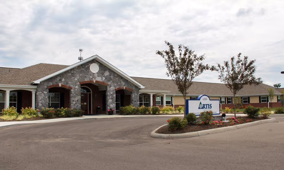 Exterior view of Artis Senior Living of Mason building with a stone and brick facade, a driveway, landscaped garden beds with flowers and small trees, and a sign displaying the facility's name.
