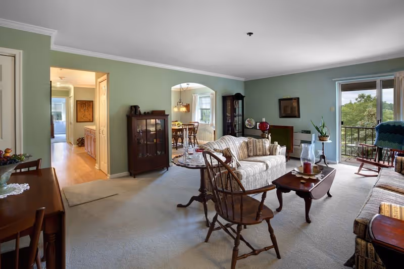 A cozy living room with light green walls and beige carpet featuring a striped sofa, wooden chairs, a coffee table with decorative items, and a glass cabinet. The room opens to a dining area with a wooden table and chairs, and a sliding glass door leads to a balcony with a view of trees outside.