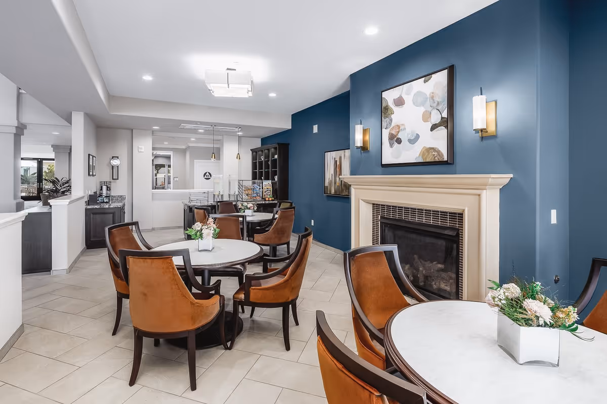 Interior view of a senior living facility common area with round tables and brown cushioned chairs. The room features a fireplace with a beige mantel on a blue accent wall, decorated with abstract paintings and wall sconces. The floor is tiled, and there are flower arrangements on the tables. In the background, there is a reception or service counter area with pendant lights and a clock on the wall.