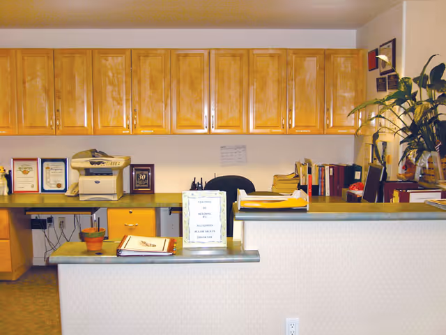 Reception desk area with a green countertop, wooden cabinets mounted on the wall behind, a printer, framed certificates, office supplies, and a potted plant on the counter. There is a sign on the desk and various files and documents organized on the right side.