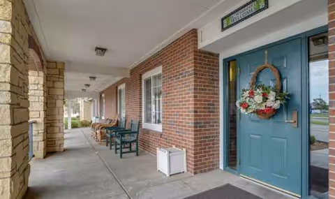 Brick building entrance with teal door, wreath, and porch seating along a covered walkway.