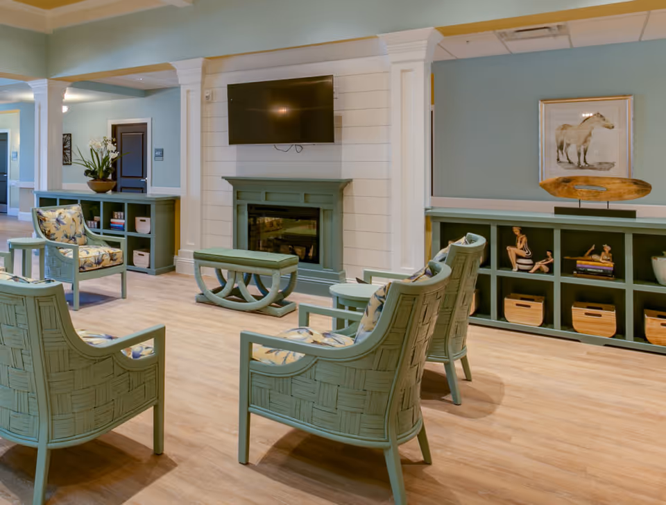 Light-filled communal lounge with mint-green woven chairs facing a fireplace topped by a wall-mounted TV.