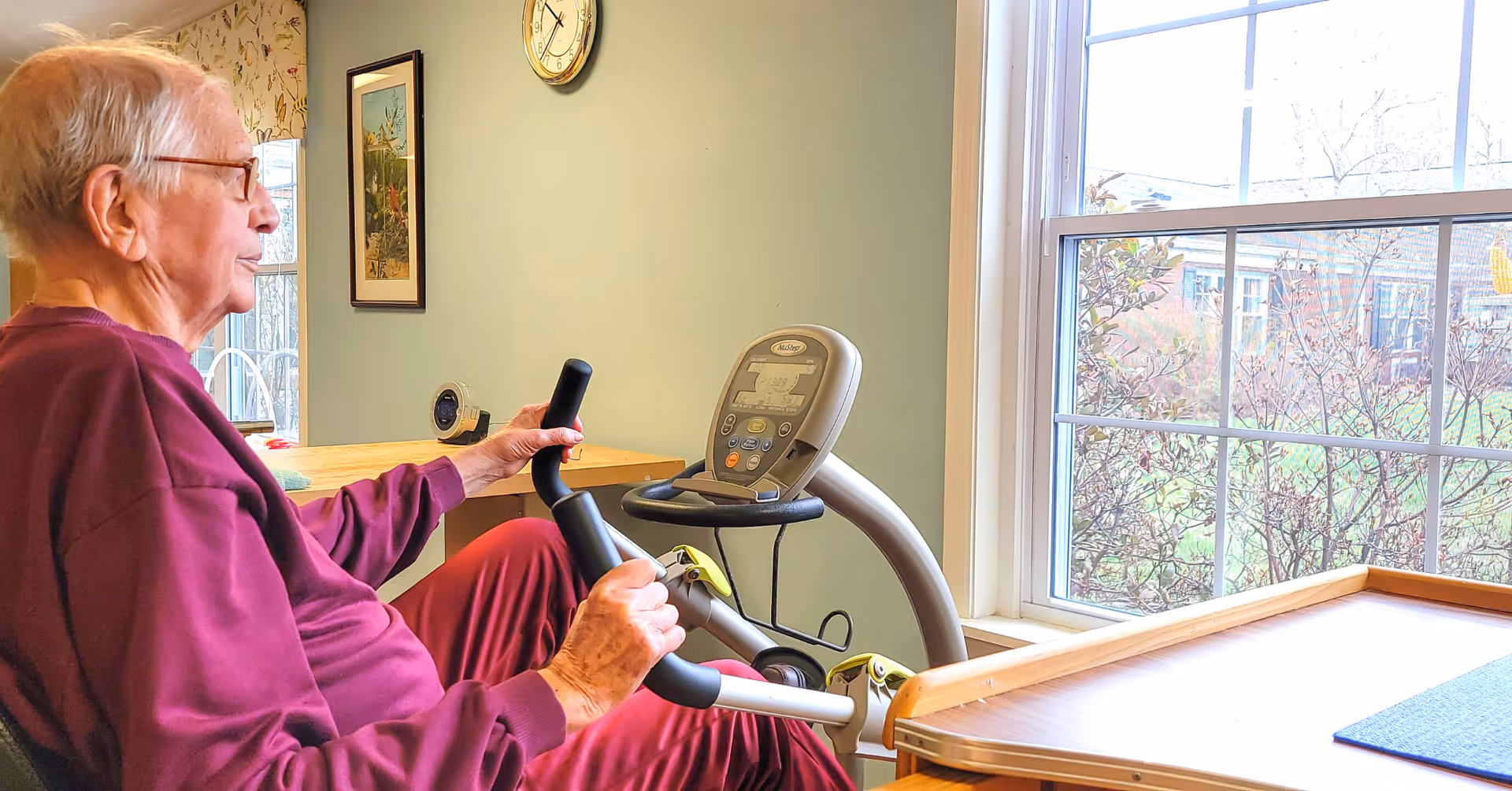 An elderly man wearing glasses and a maroon sweatshirt is seated on a stationary exercise bike indoors near a large window with a view of trees and buildings outside. The room has light green walls, a clock, and framed artwork on the wall.