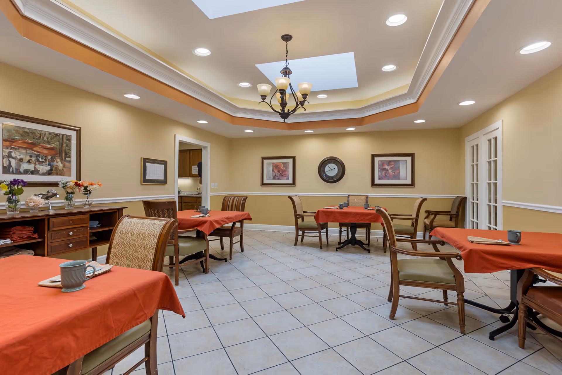 A dining room with several tables covered in red tablecloths, each set with a cup and napkin. The room has beige walls with white trim, a tiled floor, and a ceiling with recessed lighting and a chandelier. There are framed pictures and a clock on the walls, and a sideboard with flowers and decorative items.