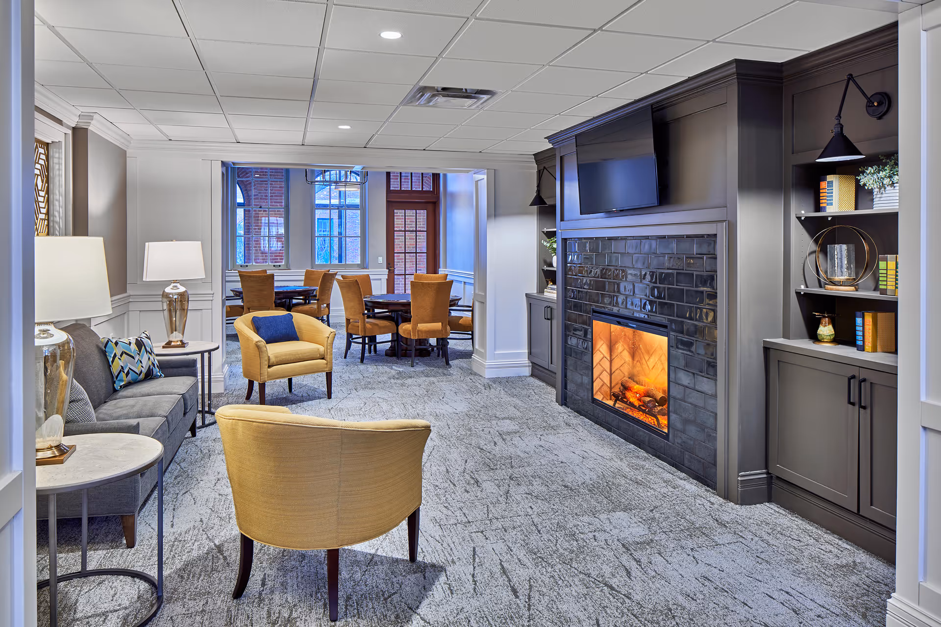A comfortable senior living lounge with armchairs and a sofa facing a fireplace and TV, with a dining table area visible in the background.