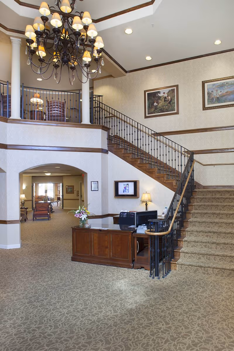 Spacious lobby with a wooden reception desk, carpeted staircase, ornate chandelier and seating area.