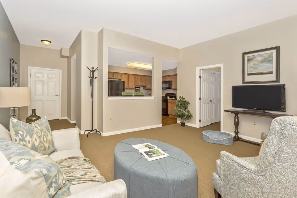 A cozy living room with beige walls and carpet. The room features a light-colored sofa with patterned pillows, a blue oval ottoman with an open book on top, and a patterned armchair. A flat-screen TV is mounted on a wooden stand against the wall, with a framed painting above it. There is a small plant near the entrance to the kitchen, which has wooden cabinets and black appliances. A coat rack stands near the front door.