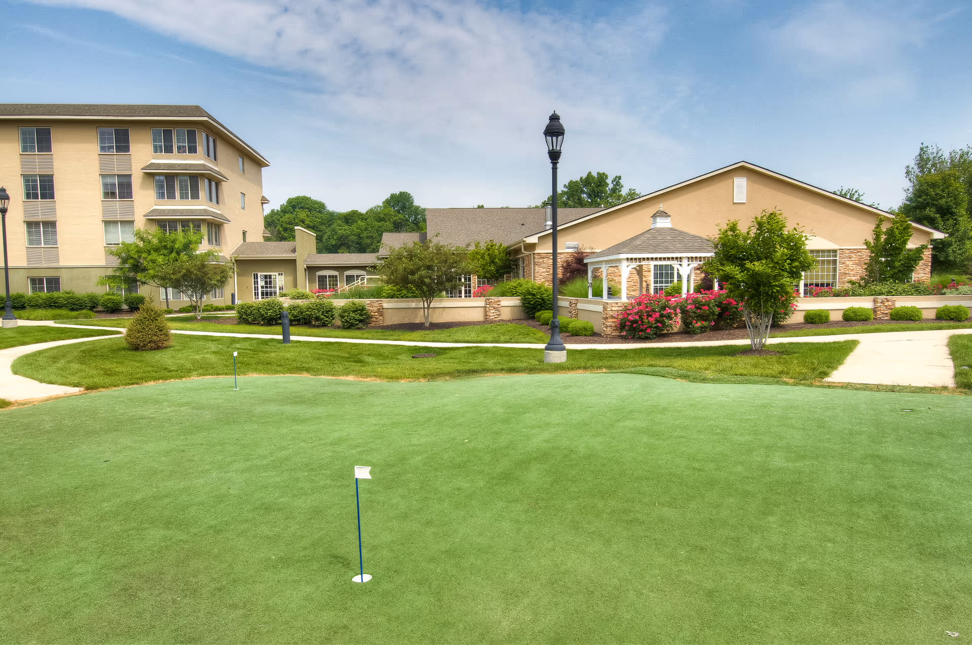 Outdoor view of Westside Garden Plaza featuring a well-maintained putting green with two small flags, surrounded by landscaped grass, bushes, and trees. In the background, there are beige buildings with multiple windows, a white gazebo, and a lamppost under a partly cloudy sky.