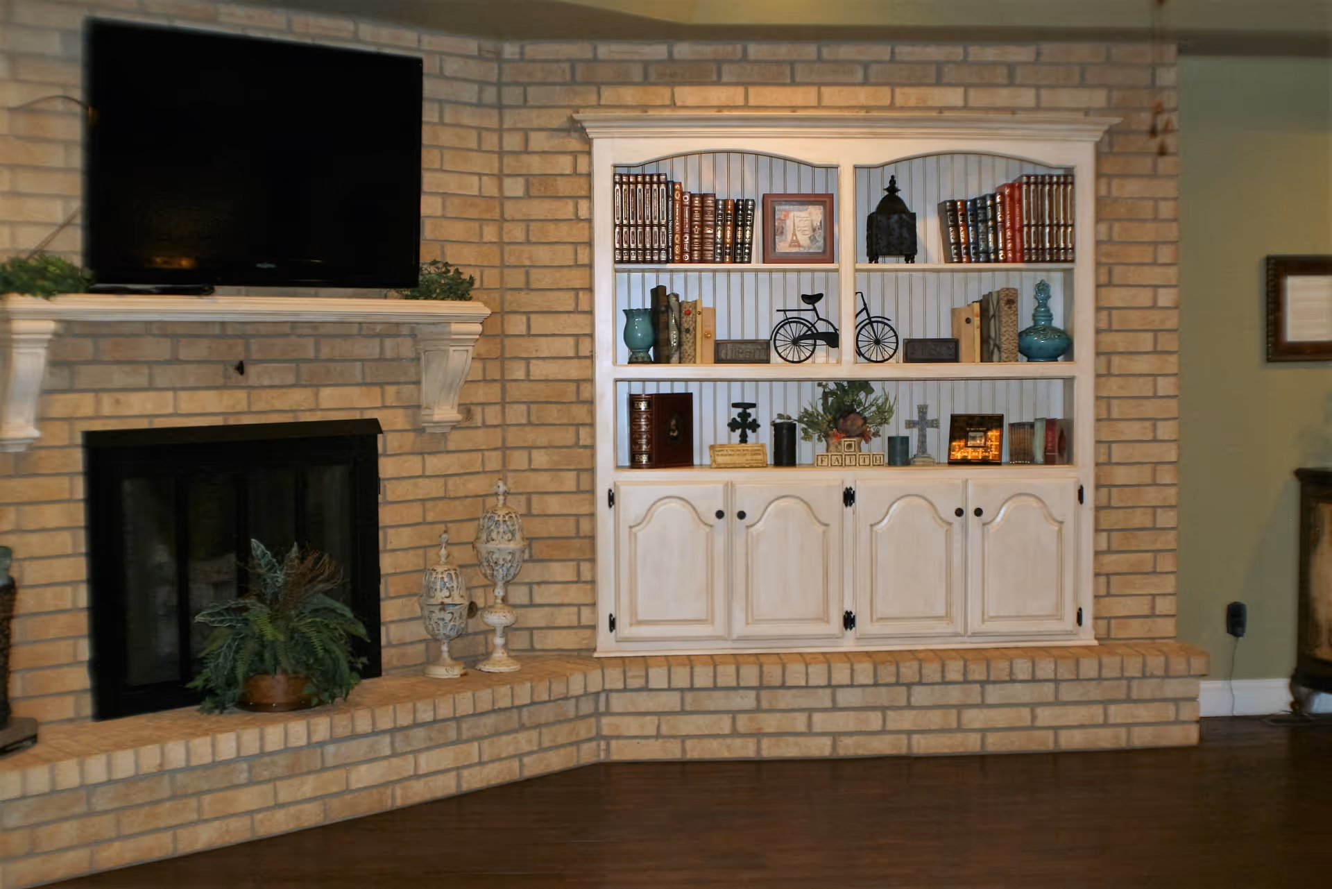 Brick fireplace with a mounted TV and a built-in white shelving unit filled with books and decorative items in a living room.