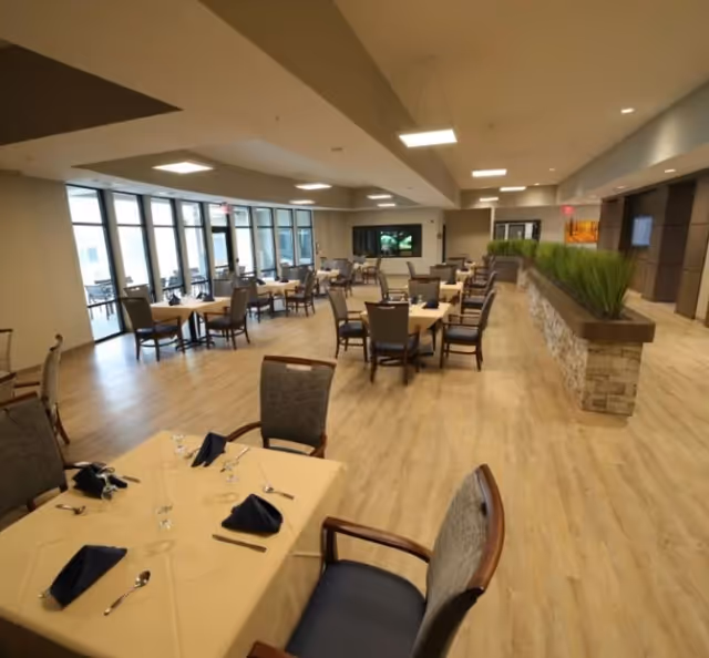 Spacious, well-lit dining room with multiple set tables and chairs, large windows along one wall, and a long planter divider.