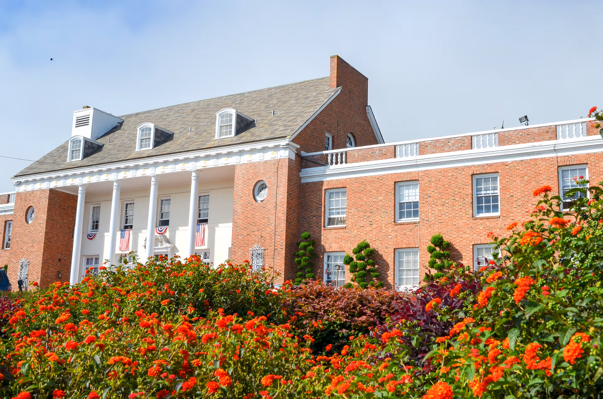 A large brick building with white columns and American flags hanging from the front porch. The building has multiple windows and a sloped roof with dormer windows. In the foreground, there is a garden filled with vibrant red and orange flowers and neatly trimmed green bushes under a clear blue sky.