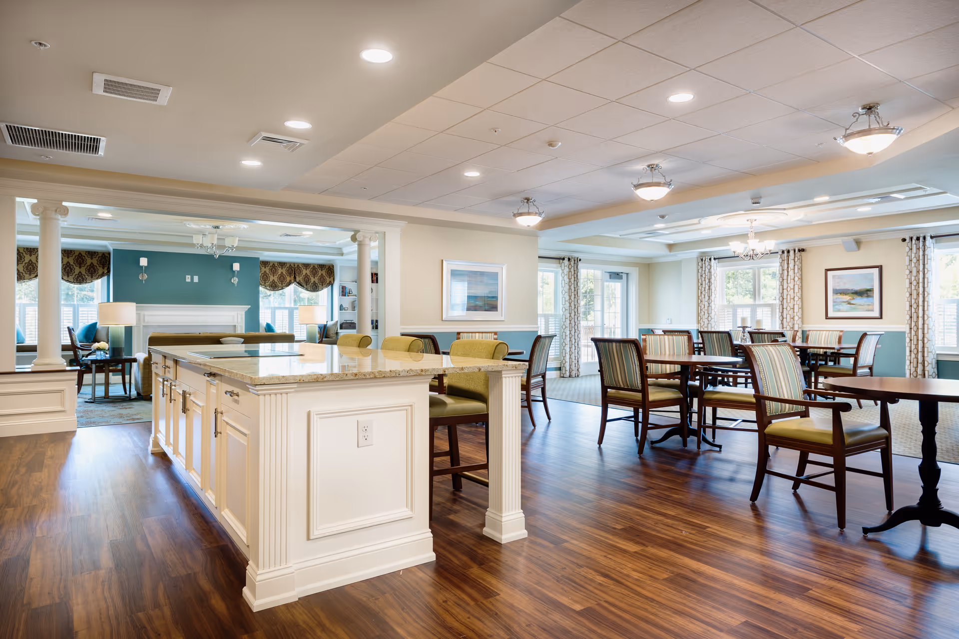 A spacious and well-lit common area in a senior living facility featuring a large kitchen island with granite countertop and several chairs. The room has wooden flooring and is furnished with multiple tables and chairs arranged for dining or socializing. In the background, there is a cozy living area with a fireplace, armchairs, and large windows with patterned valances. The walls are painted in soft tones with decorative molding, and ceiling lights provide ample illumination.