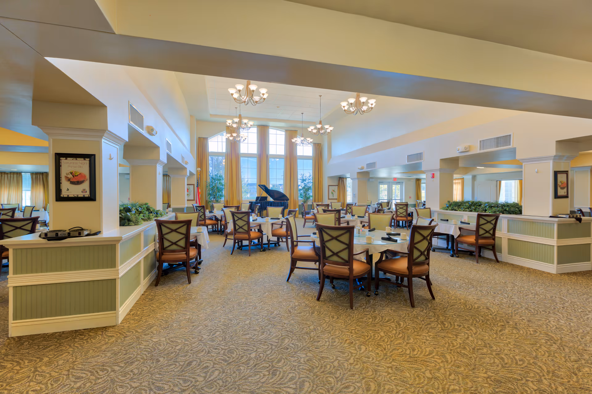 A spacious dining room with multiple round tables and chairs arranged neatly. The room features large windows with long curtains allowing natural light to fill the space. Several chandeliers hang from the ceiling, and a grand piano is positioned near the windows. The walls are light-colored with decorative columns and mirrors, and the carpet has a patterned design.