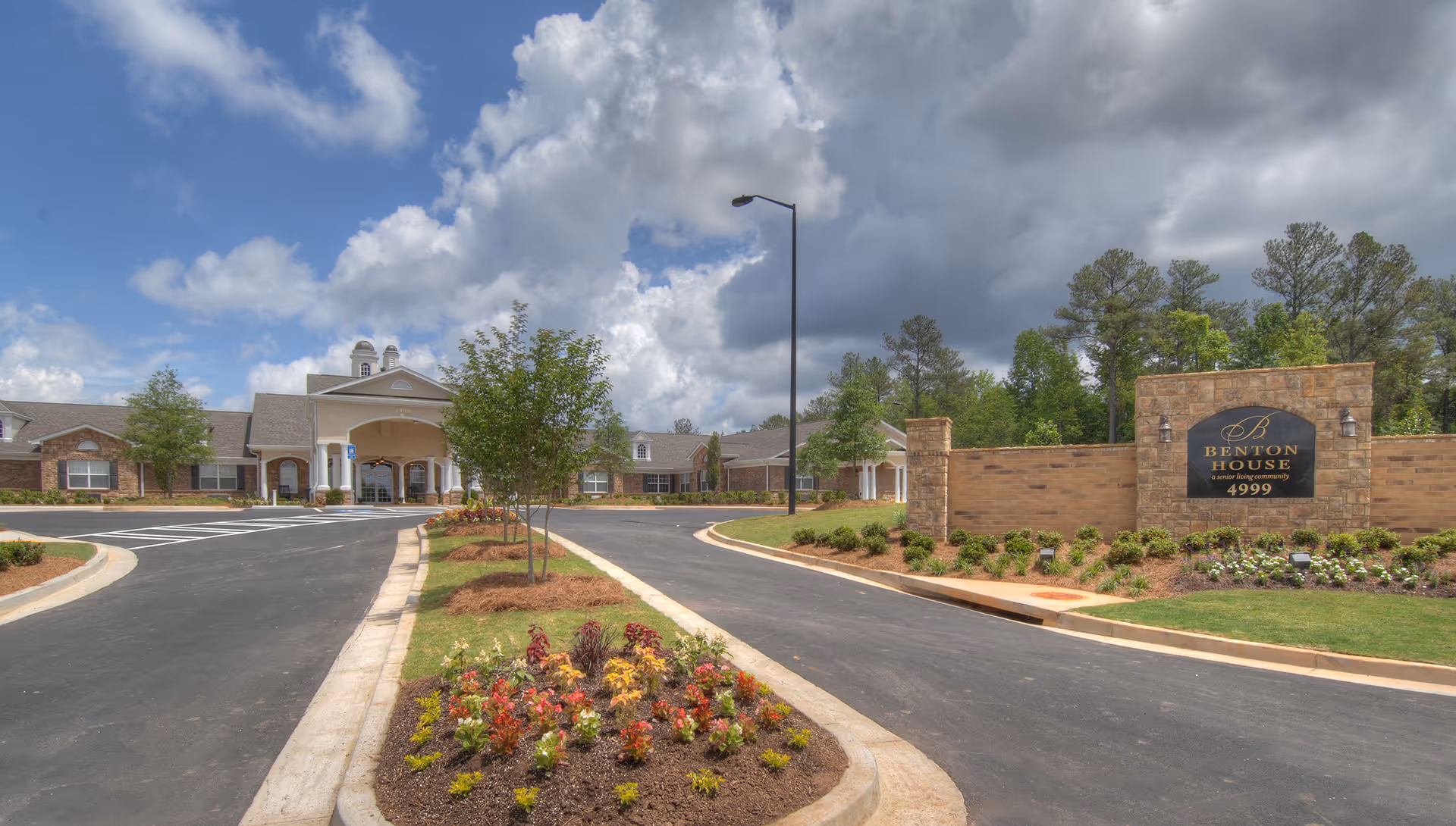Front exterior view of Benton House of Douglasville, a senior living community, showing a driveway with landscaped flower beds and trees leading to the building entrance under a partly cloudy sky.