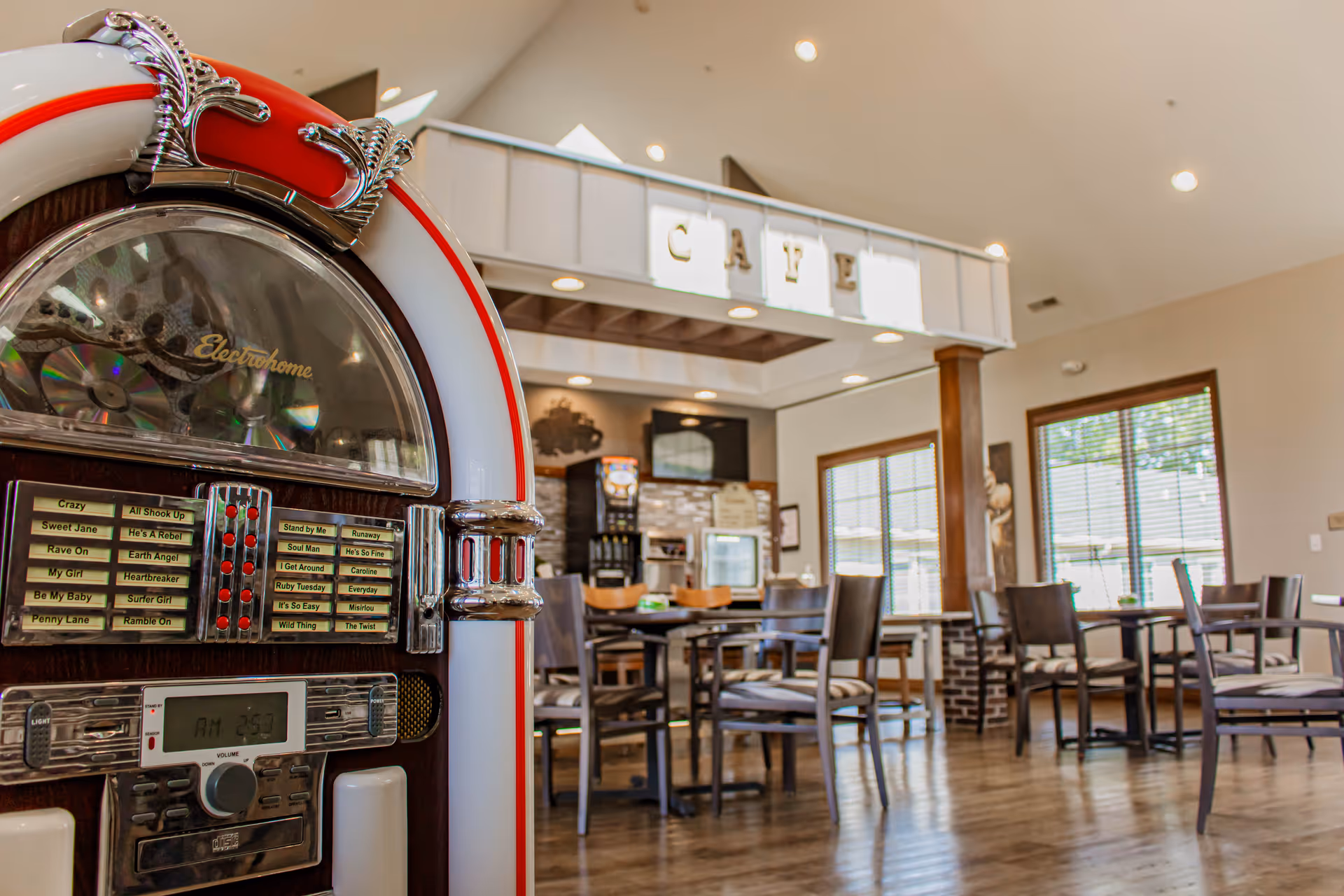 Interior view of a cafe area in an assisted living facility featuring a vintage jukebox in the foreground and several tables and chairs arranged in the background under a sign that reads 'CAFE'. Large windows allow natural light to fill the space.