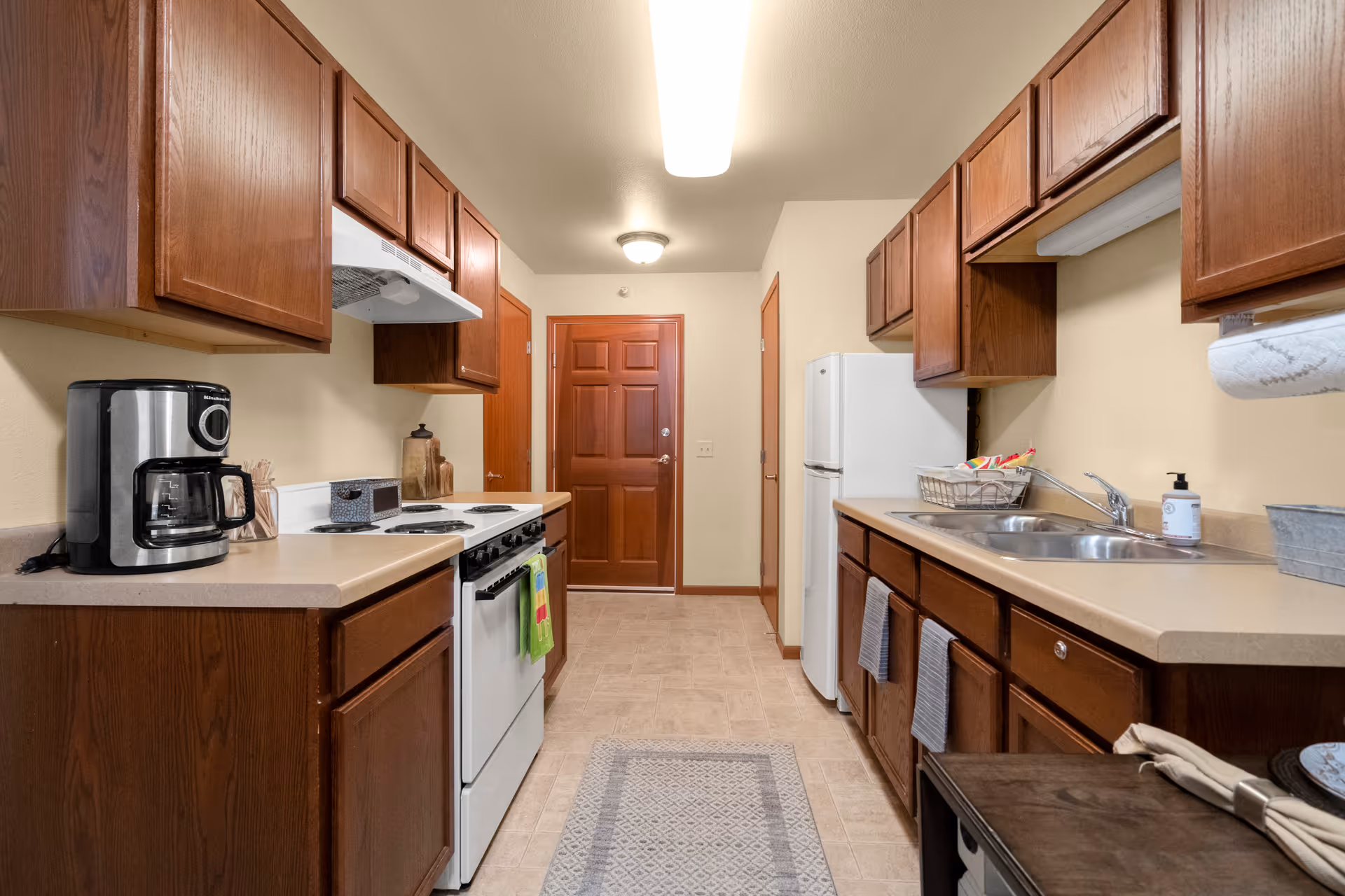 A galley-style kitchen with wooden cabinets on both sides, a white stove and oven, a white refrigerator, a coffee maker on the counter, a double sink, and a door at the end of the room. The floor has a light-colored tile and a patterned rug in the center.