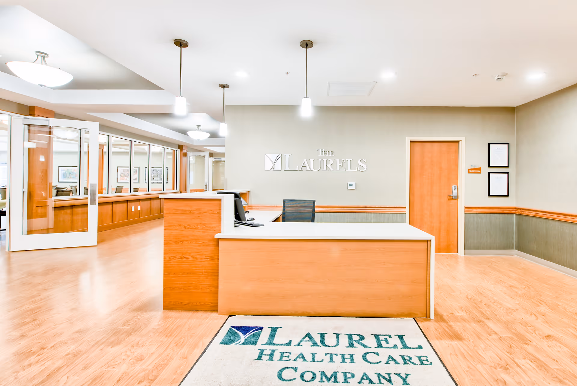 Reception area of Laurels of West Columbus featuring a wooden front desk with a computer and chair, a wall sign that reads 'The Laurels', a wooden door, framed documents on the wall, and a floor mat with the text 'Laurel Health Care Company'. The area has wood flooring, pendant lights, and glass windows along the left side.