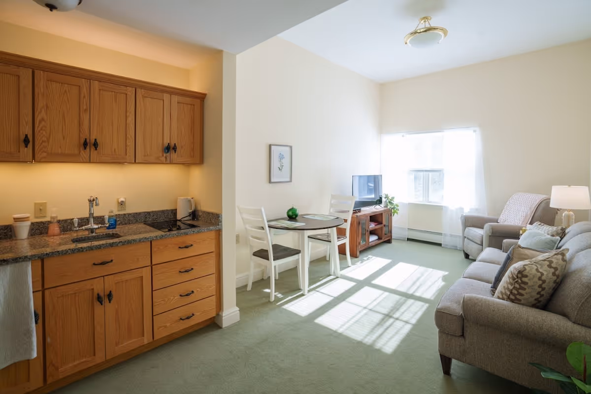 A bright and cozy living space featuring a small kitchenette with wooden cabinets and granite countertops on the left, a round dining table with two white chairs in the center, and a living area with a gray sofa, armchair, and a TV on a wooden stand near a window with sheer white curtains letting in sunlight.