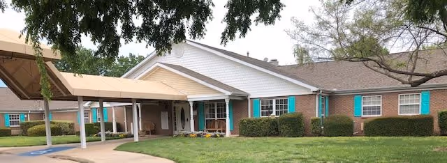 Exterior view of a single-story brick building with white siding and teal window shutters. The building has a covered entrance with a beige canopy and a well-maintained lawn with shrubs and trees in front.