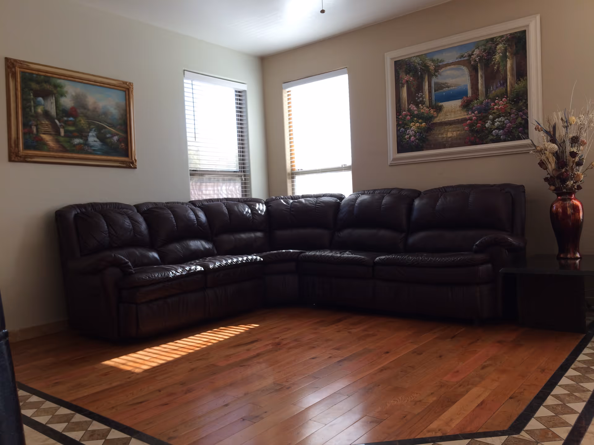 A living room with a large dark brown leather sectional sofa placed against two walls. There are two windows with blinds letting in natural light. The walls are decorated with two framed paintings depicting garden and nature scenes. A tall vase with dried flowers is placed on a small black side table next to the sofa. The floor is wooden with a decorative tile border.