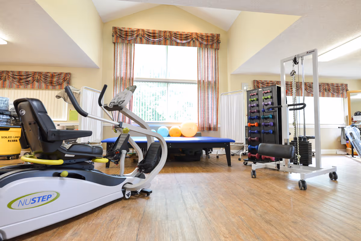A rehabilitation and wellness room with exercise equipment including a NuStep recumbent cross trainer, a weight machine, a rack of colorful dumbbells, and a blue therapy table with exercise balls in front of a large window with patterned curtains.