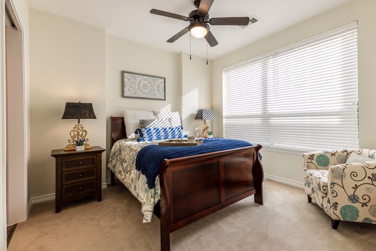 A bright and cozy bedroom with a wooden bed frame, floral bedding, and a blue throw blanket. There are two bedside tables with decorative lamps, a patterned armchair near a large window with white blinds, and a ceiling fan with a light fixture.