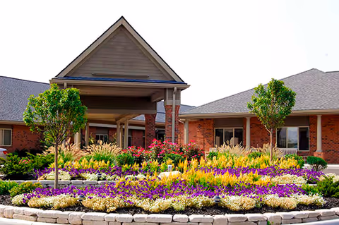 Entrance of a brick building with a covered driveway and a well-maintained garden in front featuring colorful flowers and small trees.