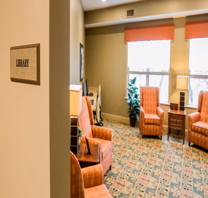 Interior view of a library room with patterned carpet, three orange upholstered armchairs, a small wooden side table with a lamp, a potted plant near the window, and a desk with a computer chair in the background. A sign on the wall near the entrance reads 'LIBRARY'.