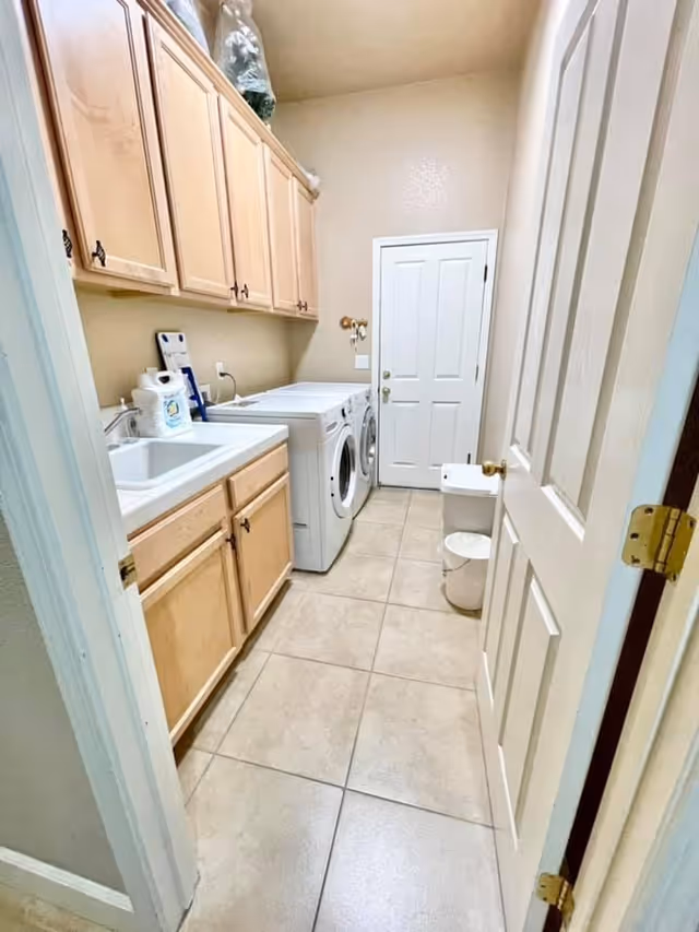 Narrow laundry room with front-loading washer and dryer, a sink, and light wood cabinets along one wall.