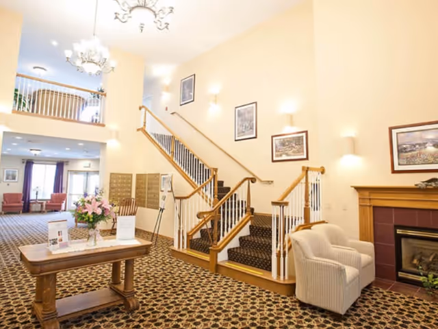 A spacious and well-lit interior common area featuring a staircase with wooden handrails and carpeted steps leading to an upper floor. The room has patterned carpet flooring, a wooden table with a flower arrangement and informational materials, a comfortable armchair next to a fireplace with a wooden mantel, and framed artwork on the walls. There is a balcony area visible above and additional seating in the background near large windows with curtains.