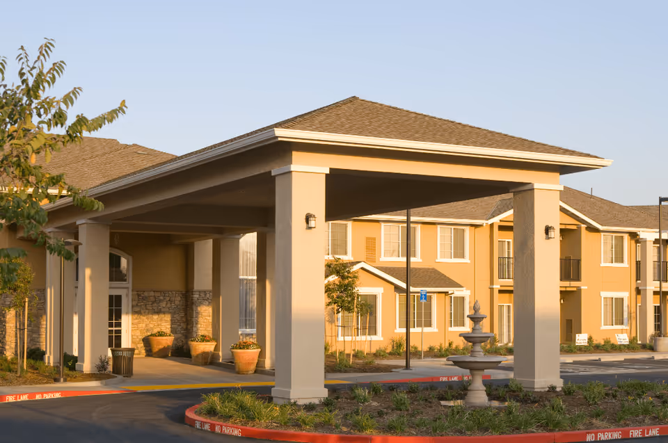 Exterior view of Manteca Manor senior living facility showing the entrance with a covered drop-off area supported by columns, landscaped greenery, and a multi-story building with windows and balconies in the background under a clear sky.