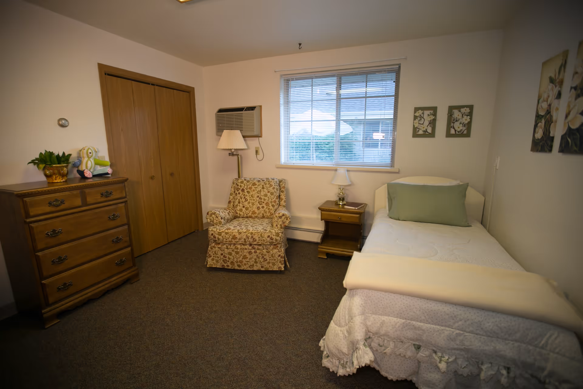 A cozy senior living bedroom with a single bed covered in white bedding and a green pillow, a wooden nightstand with a small lamp, a floral patterned armchair, a wooden dresser with a plant and a stuffed owl toy on top, a window with blinds, and floral wall art.