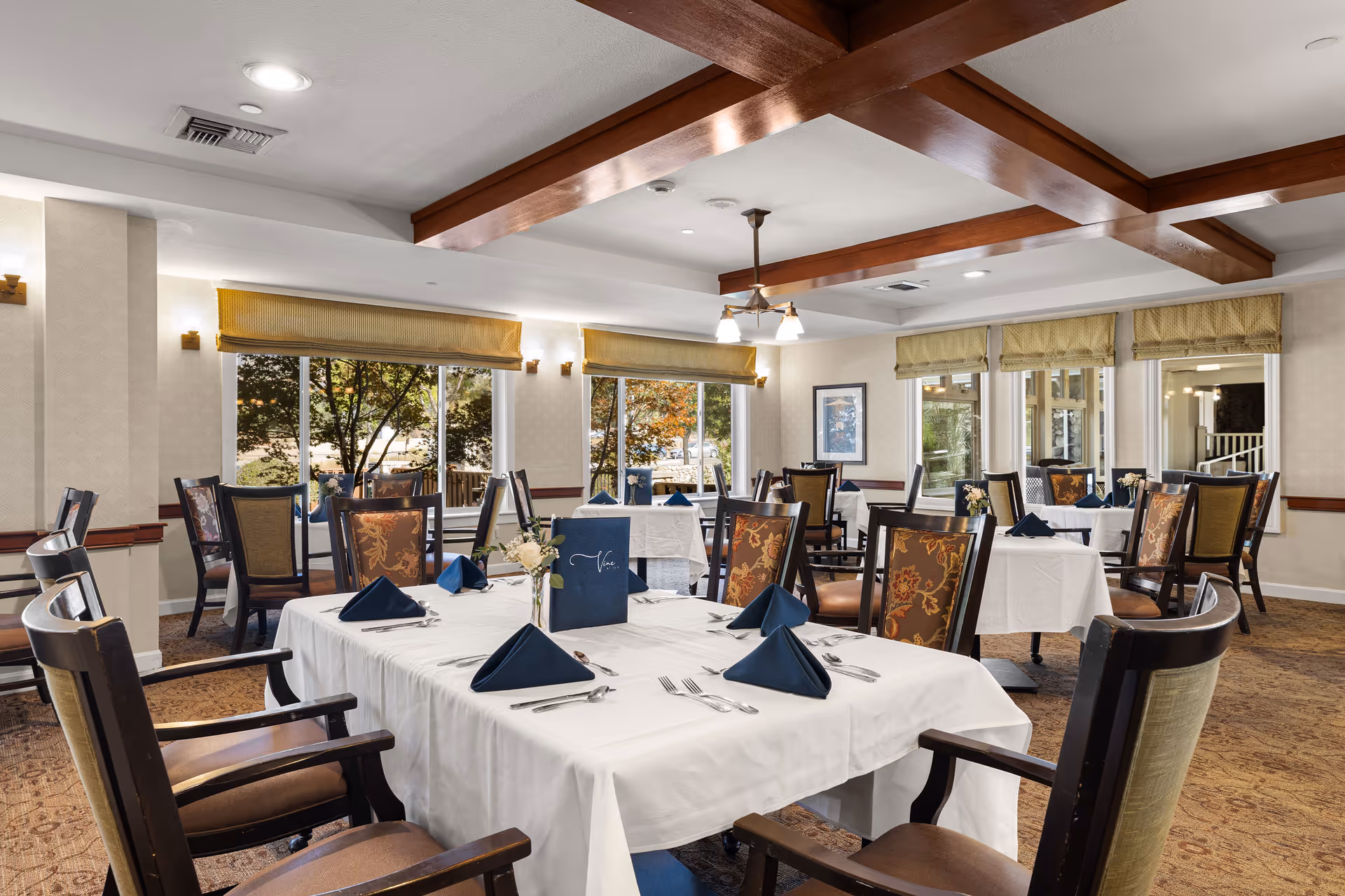 A well-lit dining room with multiple tables covered in white tablecloths, each set with silverware, folded navy blue napkins, and small floral centerpieces. The chairs have wooden frames with patterned upholstery. Large windows with yellow valances allow natural light to enter, and wooden beams adorn the ceiling.