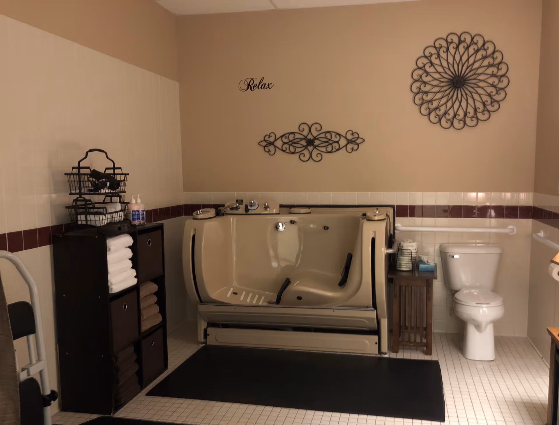A bathroom with a beige walk-in bathtub, a white toilet, and a small wooden side table holding toiletries. There is a black shelving unit with towels and baskets on the left side. The walls are tiled halfway up with white tiles and a maroon border, and the upper walls are painted beige. Decorative metal wall art and the word 'Relax' are displayed above the bathtub.